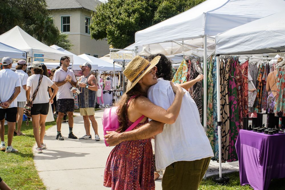 Two people embracing in the aisle between white vendor tents at Coco Market, with clothing racks and shoppers around them.